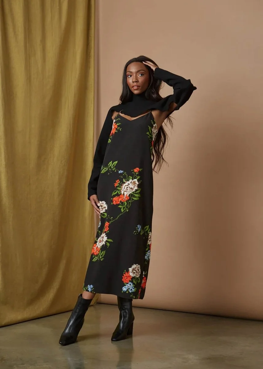 A woman standing in front of a beige wall and gold curtain, wearing a black floral dress with long sleeves, high heels, and lightly styled hair.
