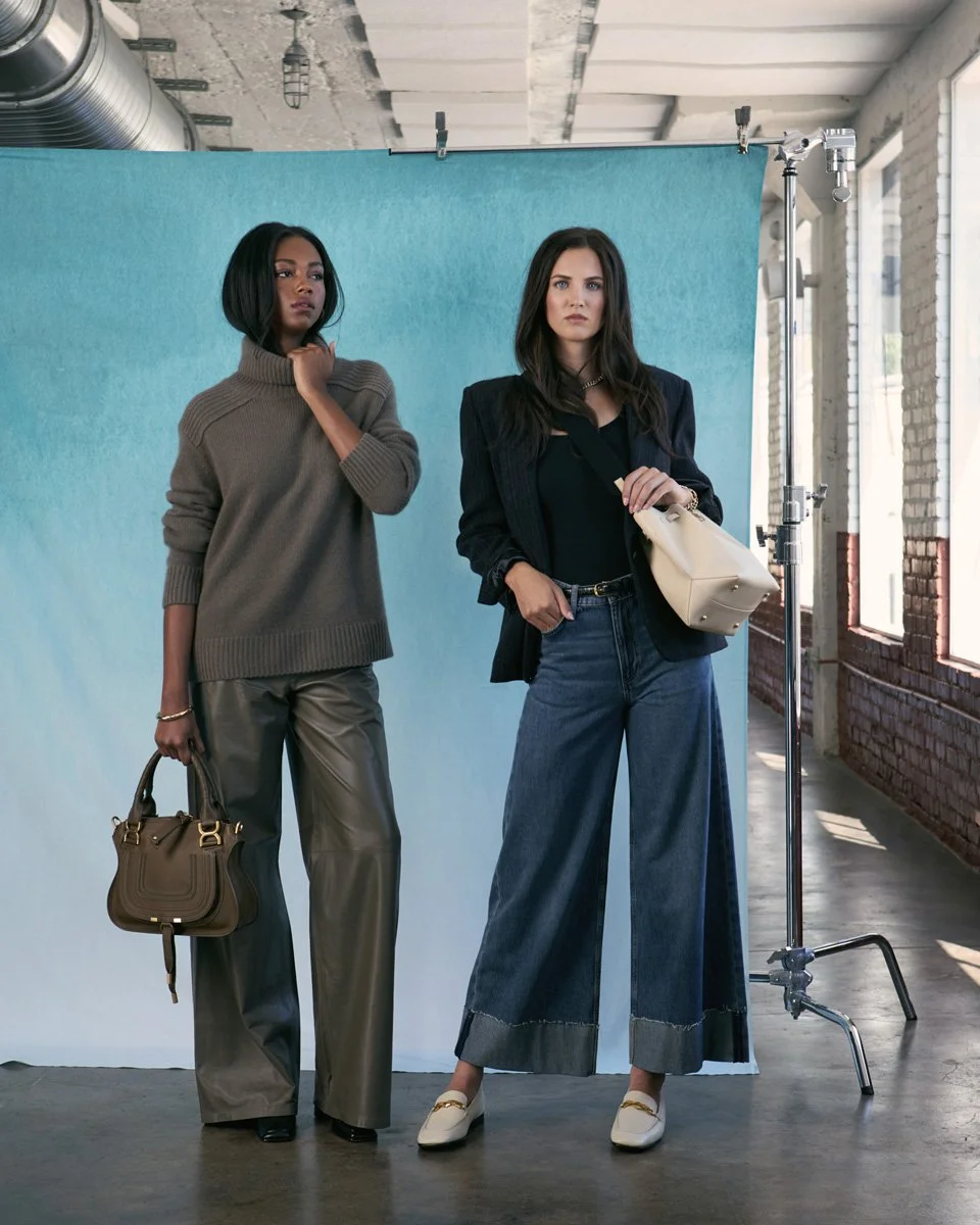 Two women standing in front of a blue backdrop in a photo studio with natural light coming through windows, dressed in stylish casual outfits.