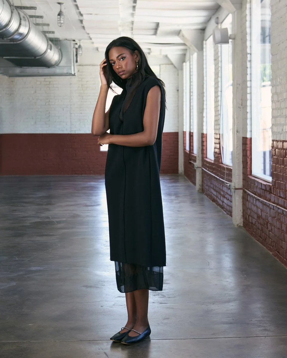 A woman standing in an industrial-style room with white brick walls and large windows, wearing a black sleeveless dress and black shoes.