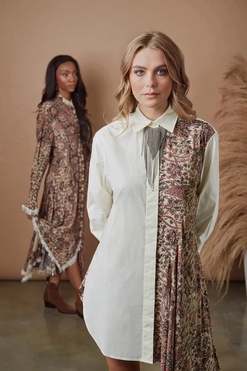 Two women standing indoors against a tan background; one in the foreground with blonde hair wearing a cream and patterned dress, the other in the background with dark hair wearing a similar patterned dress and brown boots.