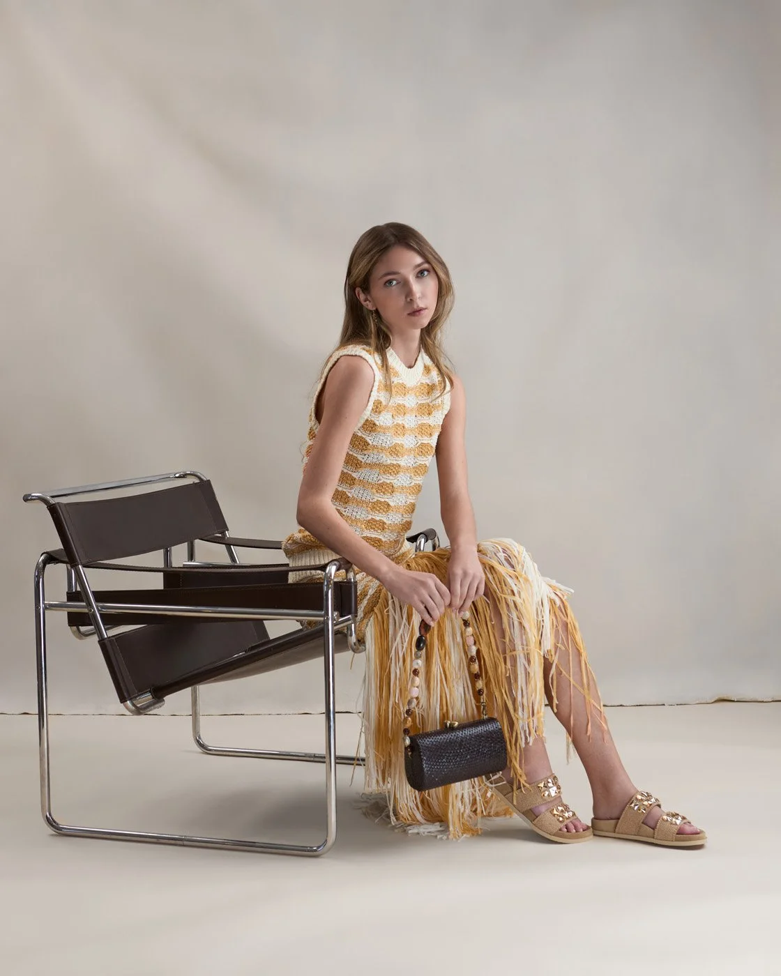 Two women posing indoors in front of a beige fabric backdrop and stone wall, surrounded by large green plants. The woman on the left has light skin, brown hair, and is wearing a dark patterned blouse, high-waisted dark green pants with gold buttons, beige ankle boots, and holding a yellow purse. The woman on the right has dark skin, black hair, and is wearing a long-sleeved, high-neck, black dress with a subtle pattern and dark heels.