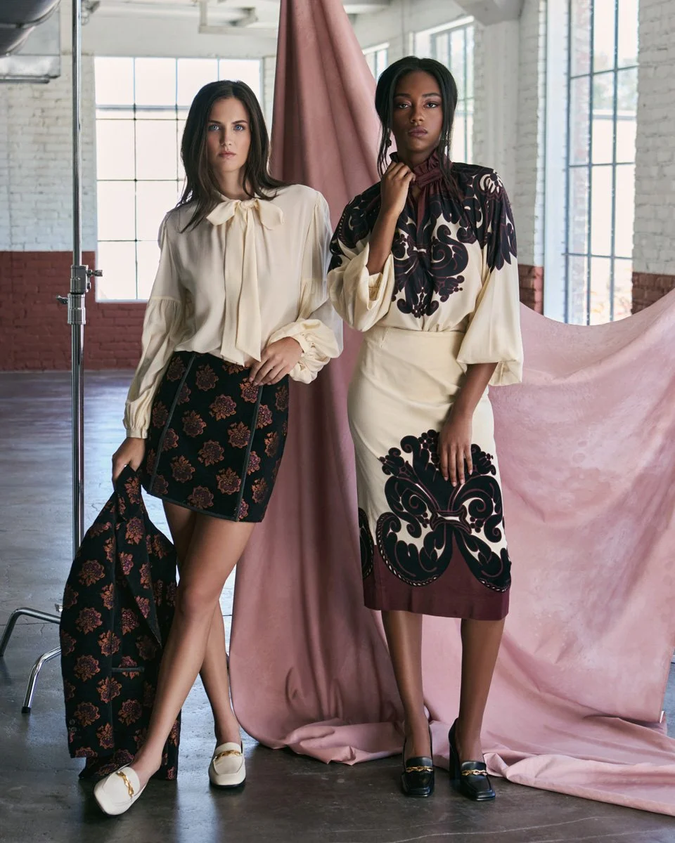 Two women posing in a studio with large windows and white brick walls. One woman is wearing a cream blouse and a patterned skirt, holding a matching coat. The other woman is dressed in a cream and dark patterned blouse and a beige skirt with black fl