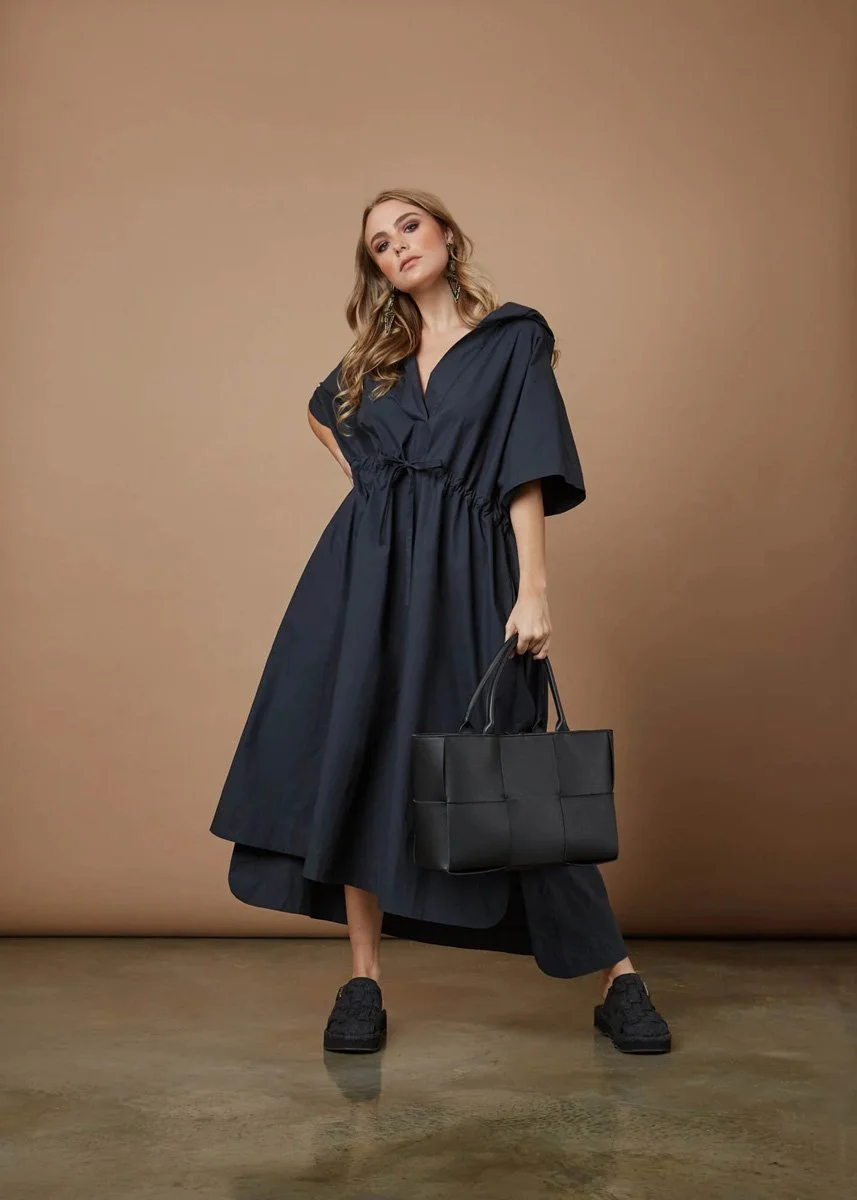 A woman with blonde wavy hair wearing a dark, loose-fitting dress or cover-up, black shoes, and holding a black handbag, standing against a plain beige background.