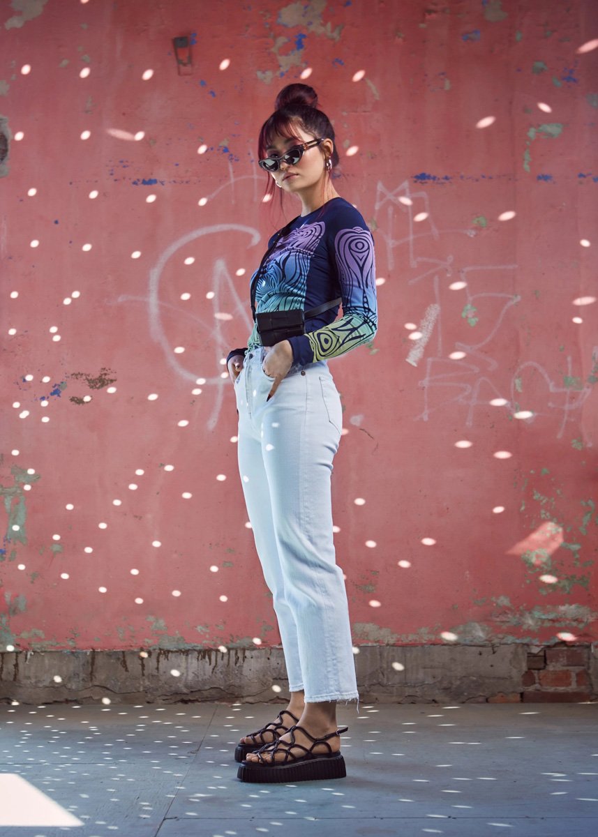 A young woman with sunglasses and a top with a colorful, abstract pattern, white high-waisted jeans, and black sandals stands against a red, graffiti-covered wall.
