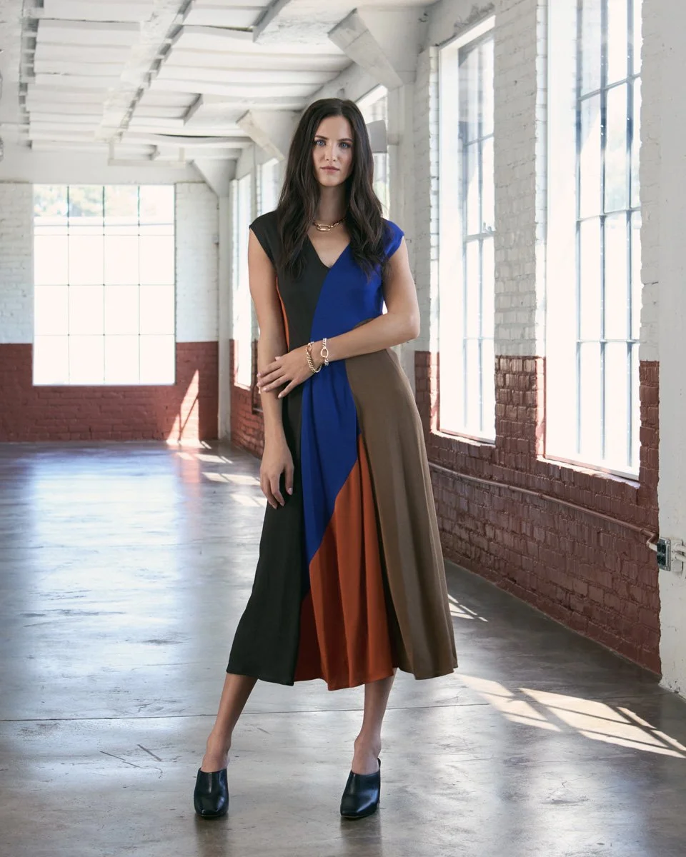A woman in a multicolored dress standing inside a spacious industrial-style loft with large windows and brick walls.