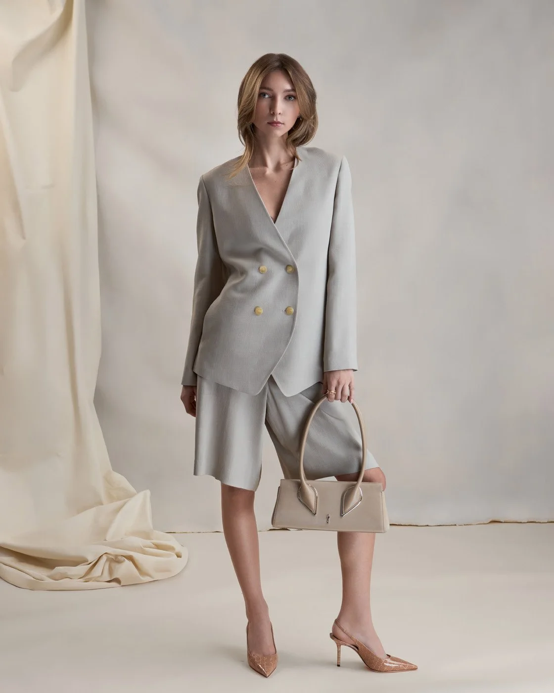 Two women posing indoors with a stone wall and a beige fabric backdrop behind them. One woman with long dark hair, wearing a beige sweater, black leather pants, and black heeled boots, sitting on a wooden chair. The other woman with long wavy brown hair, wearing a dark denim shirt, a tan cardigan draped over her shoulders, dark jeans, and tan heeled boots, standing beside her.