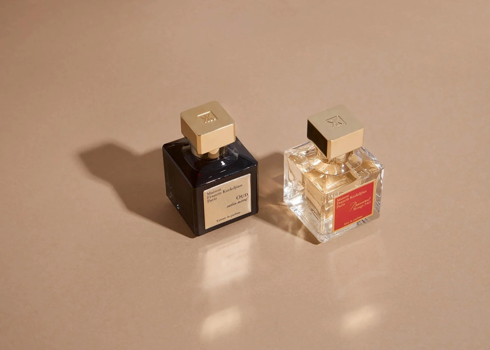 Two perfume bottles on a beige surface, one with dark liquid and the other with clear liquid, both with gold caps and labels.