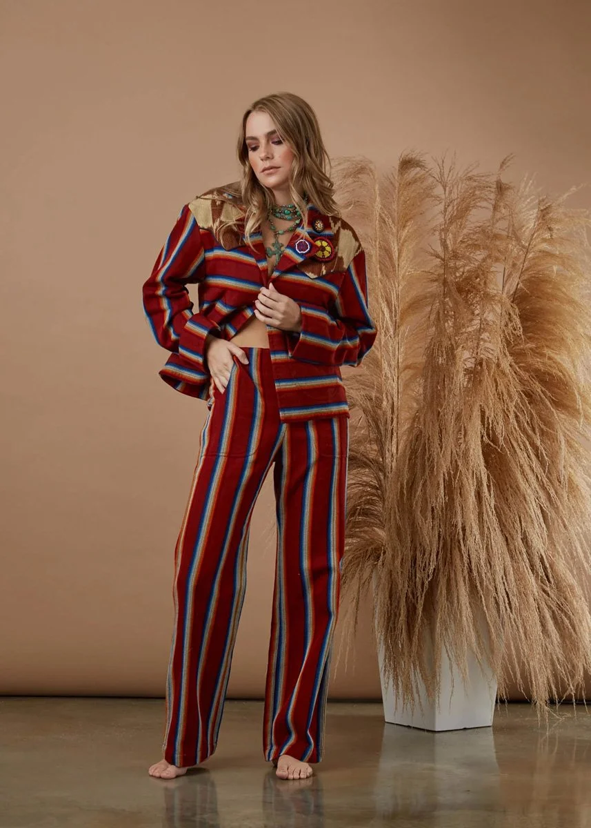 A woman in a colorful striped suit standing barefoot indoors next to a large textured plant with dried grass in a white pot.