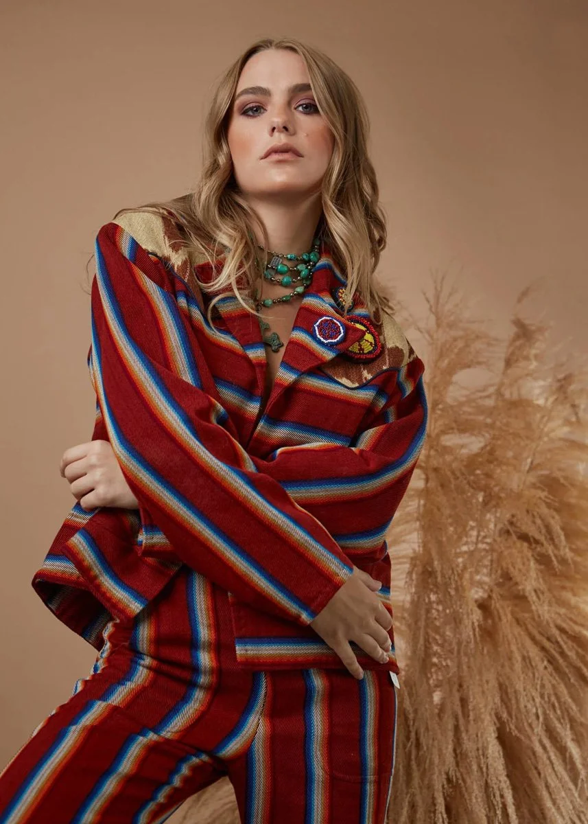 A woman in a colorful, striped jacket and matching pants, accessorized with turquoise jewelry, standing against a neutral background with dried grasses in the background.