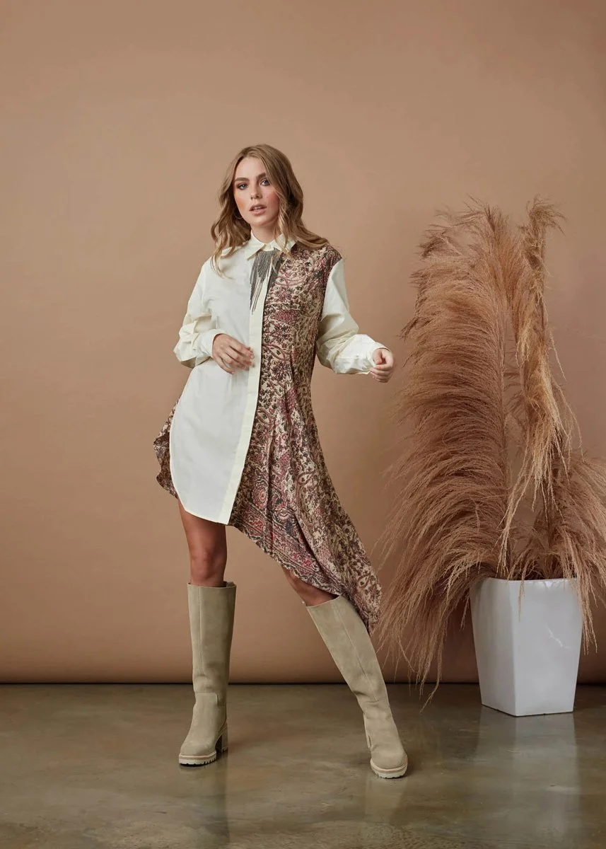 A woman posing indoors in a mixed outfit, with beige knee-high boots, a long shirt with a paisley pattern on the right side, and a white shirt underneath, standing next to a large vase filled with pampas grass.