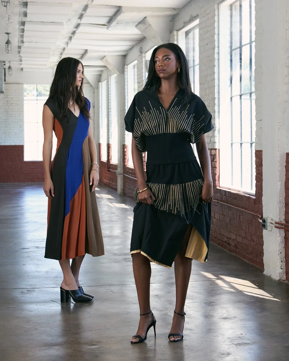 Two women standing in a bright industrial space with large windows. The woman in the foreground wears a black dress with gold detailing and high heels. The woman in the background wears a multicolored, color-block dress and black heels.