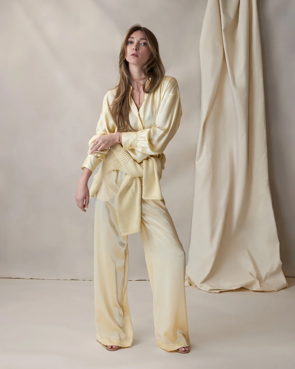 A woman with dark hair and dark skin wearing a plaid skirt and matching scarf on a light green background, with tropical leaves partially visible on the left and right sides.