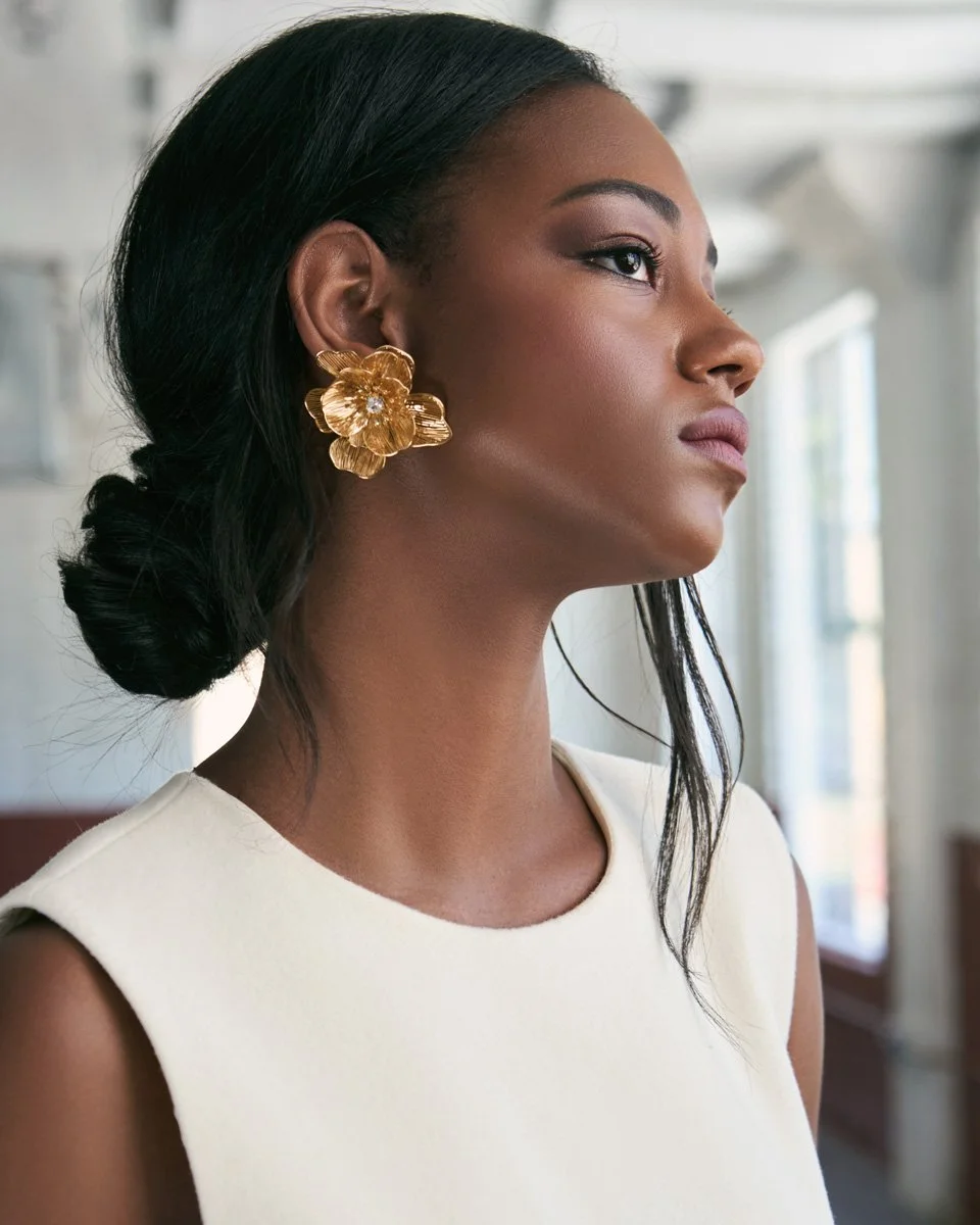 A woman with dark hair in a low bun, wearing large gold floral earrings and a sleeveless cream-colored top, standing near large windows with bright natural light.