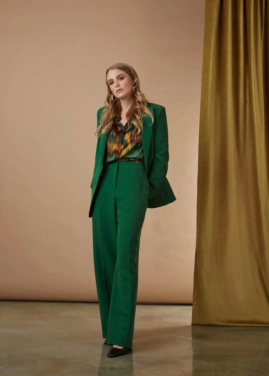 Fashion model wearing a green pantsuit with a colorful blouse, standing in front of a beige background with gold curtains.
