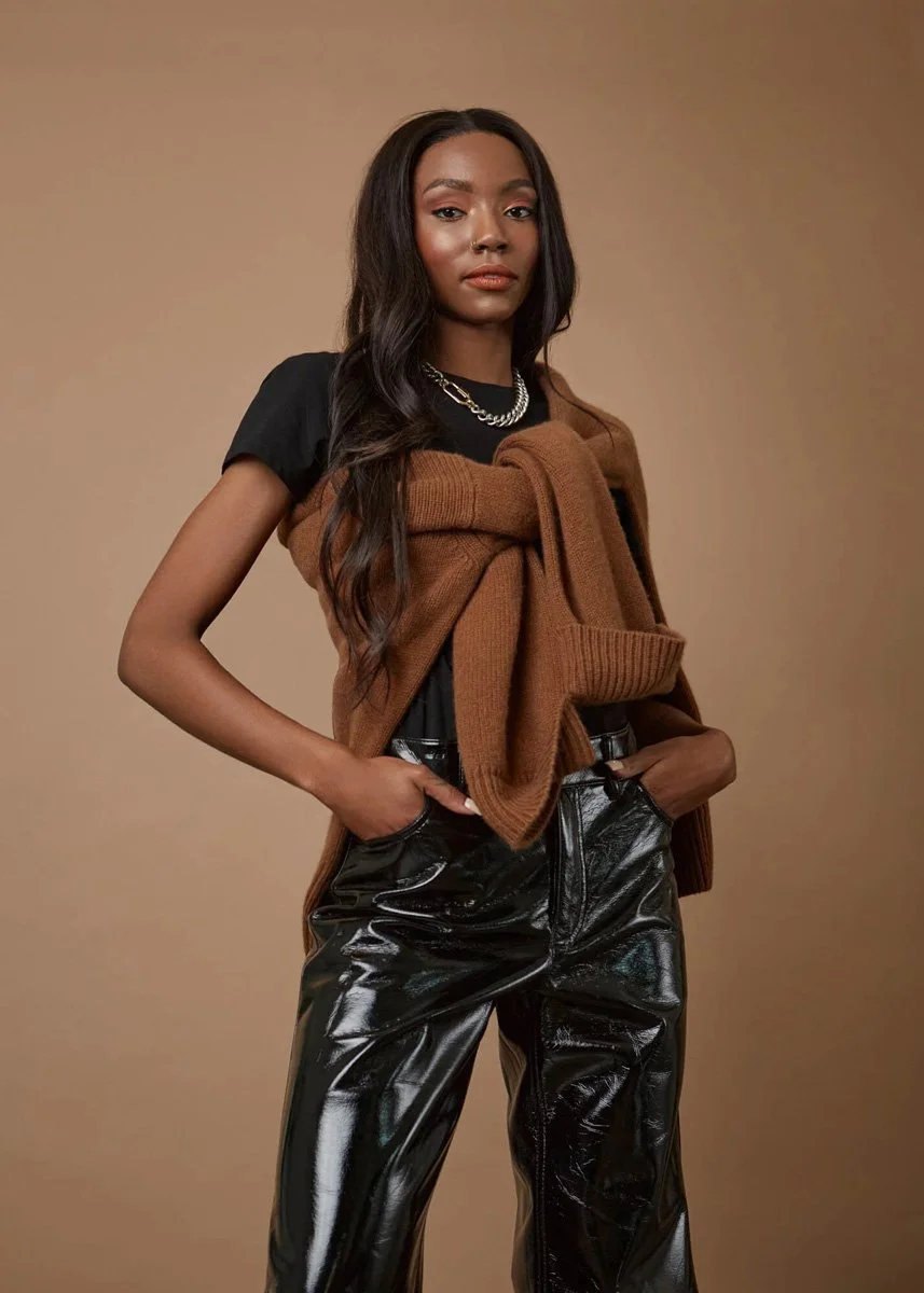 A young woman with long, wavy dark hair, wearing a black t-shirt, a brown sweater draped over her shoulders, shiny black vinyl pants, and a silver chain necklace, standing with hands in pockets against a plain beige background.