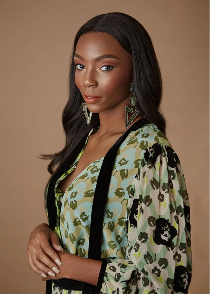 A woman with dark hair, wearing large triangular earrings, a floral-patterned jacket with black trim, and natural makeup, posing against a plain background.
