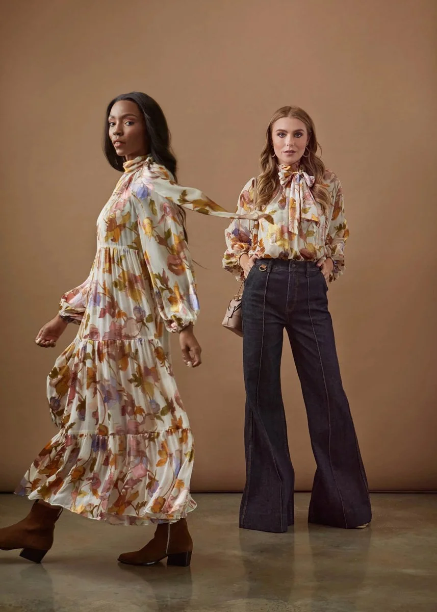 Two women standing against a beige backdrop, wearing floral patterned tops and pants, with one woman in a tiered maxi dress and the other in high-waisted wide-leg jeans.
