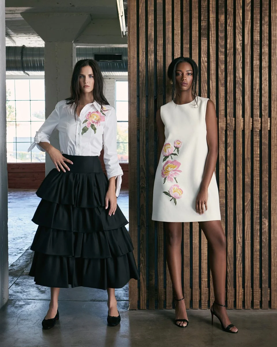Two women posing indoors, both wearing dresses with floral embroidery. The woman on the left is in a white button-down shirt with rolled sleeves and a layered black skirt, while the woman on the right wears a sleeveless white dress with pink and yell