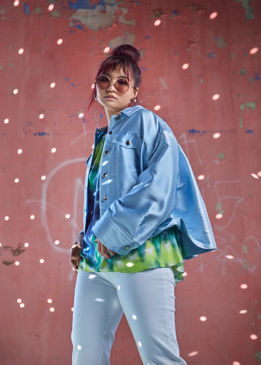 A young woman with reddish hair in a bun, wearing large round sunglasses, a shiny blue jacket, a colorful tie-dye shirt, and white pants, standing against a weathered pink wall with graffiti and surrounded by floating light spots.
