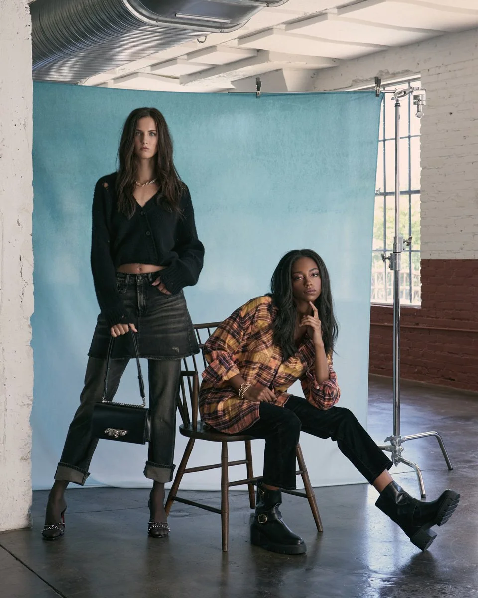 Two women in fashion poses in a studio with a light blue backdrop, one standing holding a black handbag, the other sitting on a wooden chair with her hand on her chin, wearing casual and edgy clothing.