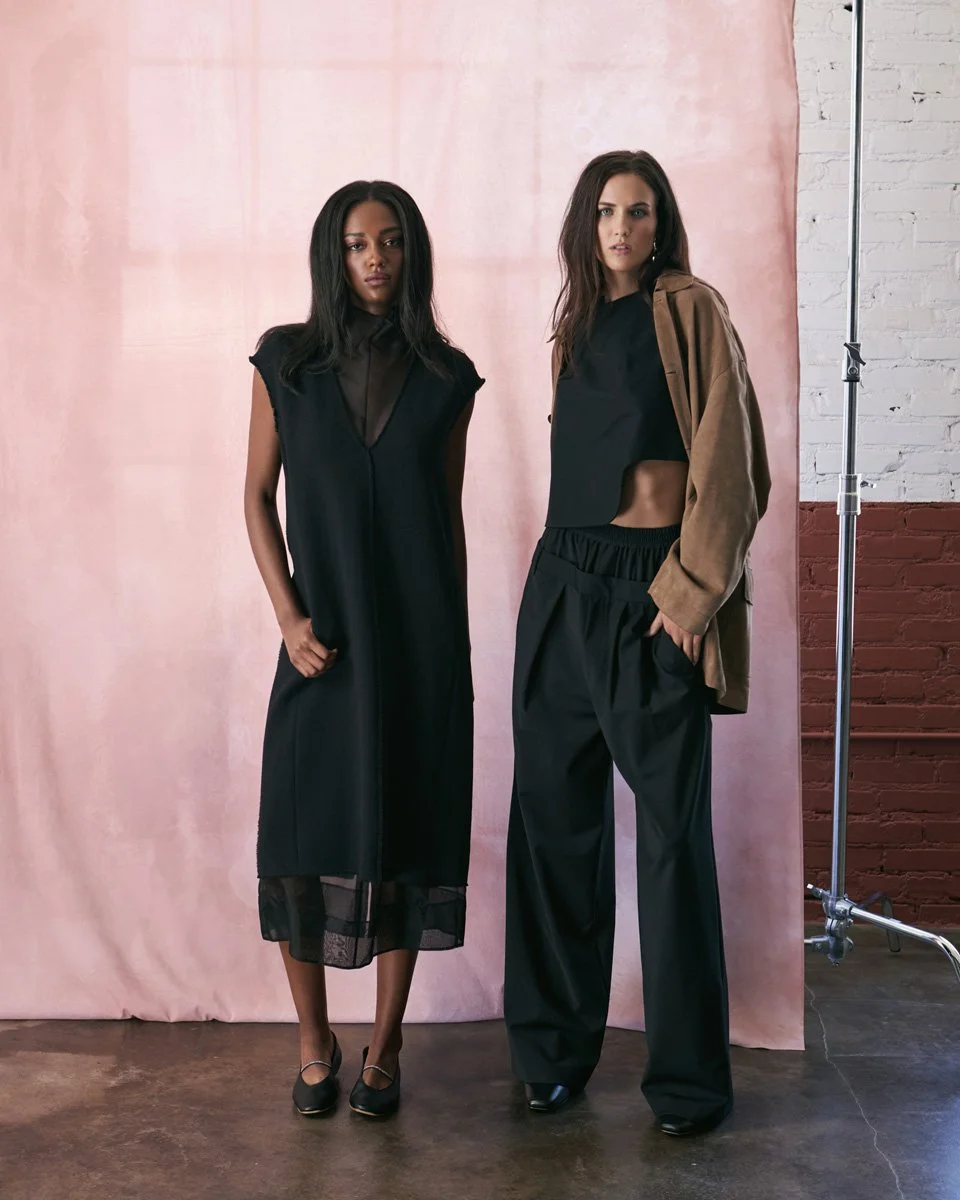Two women standing together against a pink backdrop and a white brick wall; one in a black sleeveless dress and the other in black pants with a brown jacket.