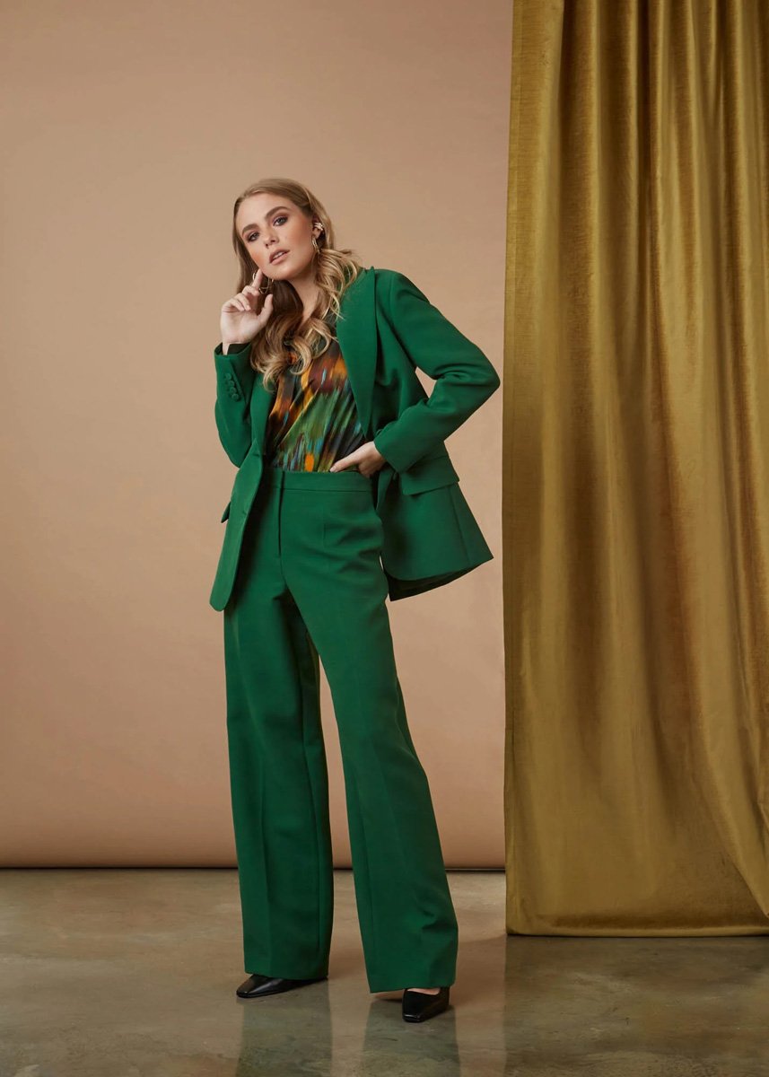 A woman with styled hair and makeup wearing a green pantsuit, black heels, and a colorful patterned blouse standing in front of a beige backdrop and gold curtain.