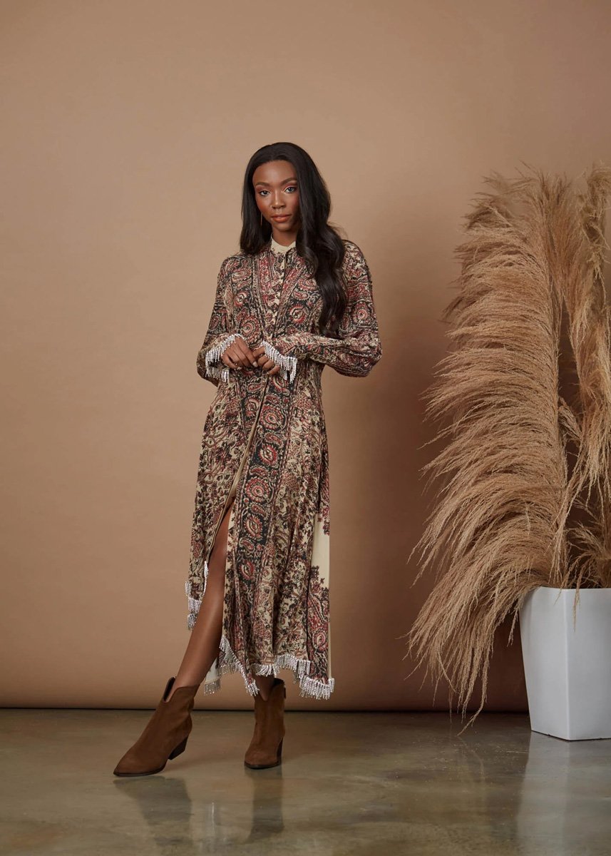 A woman standing in front of a beige background next to a large white planter filled with pampas grass, wearing a long patterned dress with a front slit and brown ankle boots.