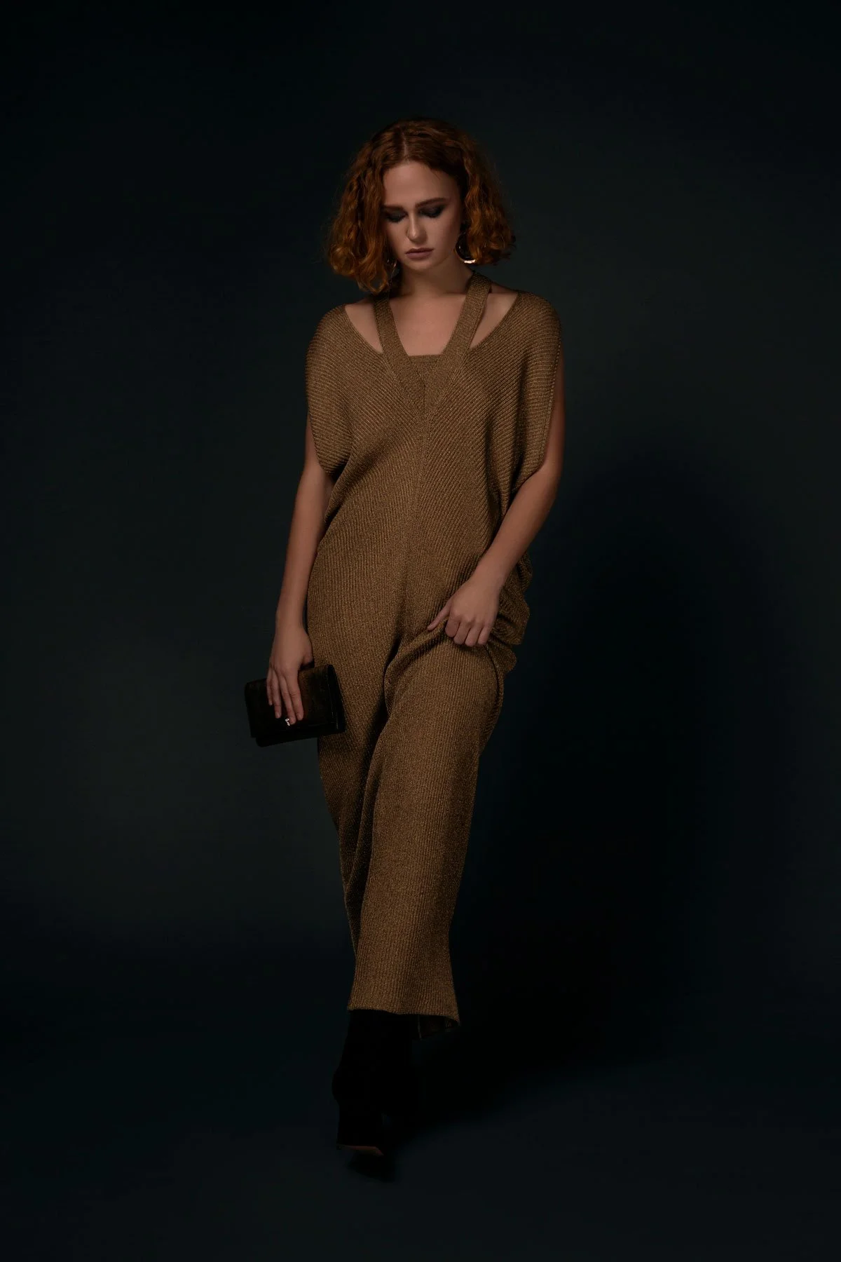 A woman with curly red hair wearing a loose, brown sleeveless jumpsuit, holding a small black clutch, posed against a dark background.