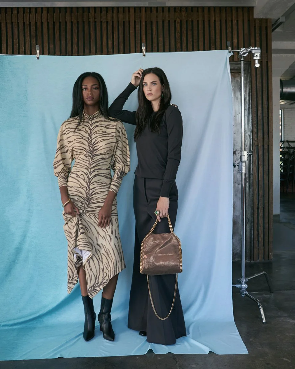 Two women standing in front of a light blue backdrop, one wearing a zebra-print dress with black boots, and the other dressed in black holding a brown bag with a gold chain.