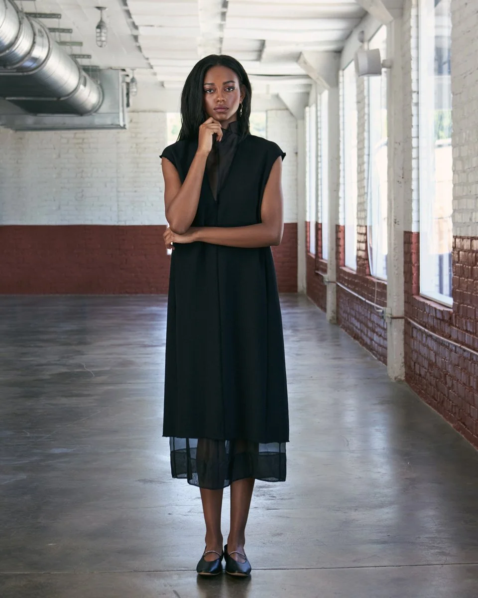 A woman stands in an industrial-style room with white brick walls and large windows, wearing a black dress and black shoes, with a pensive expression.
