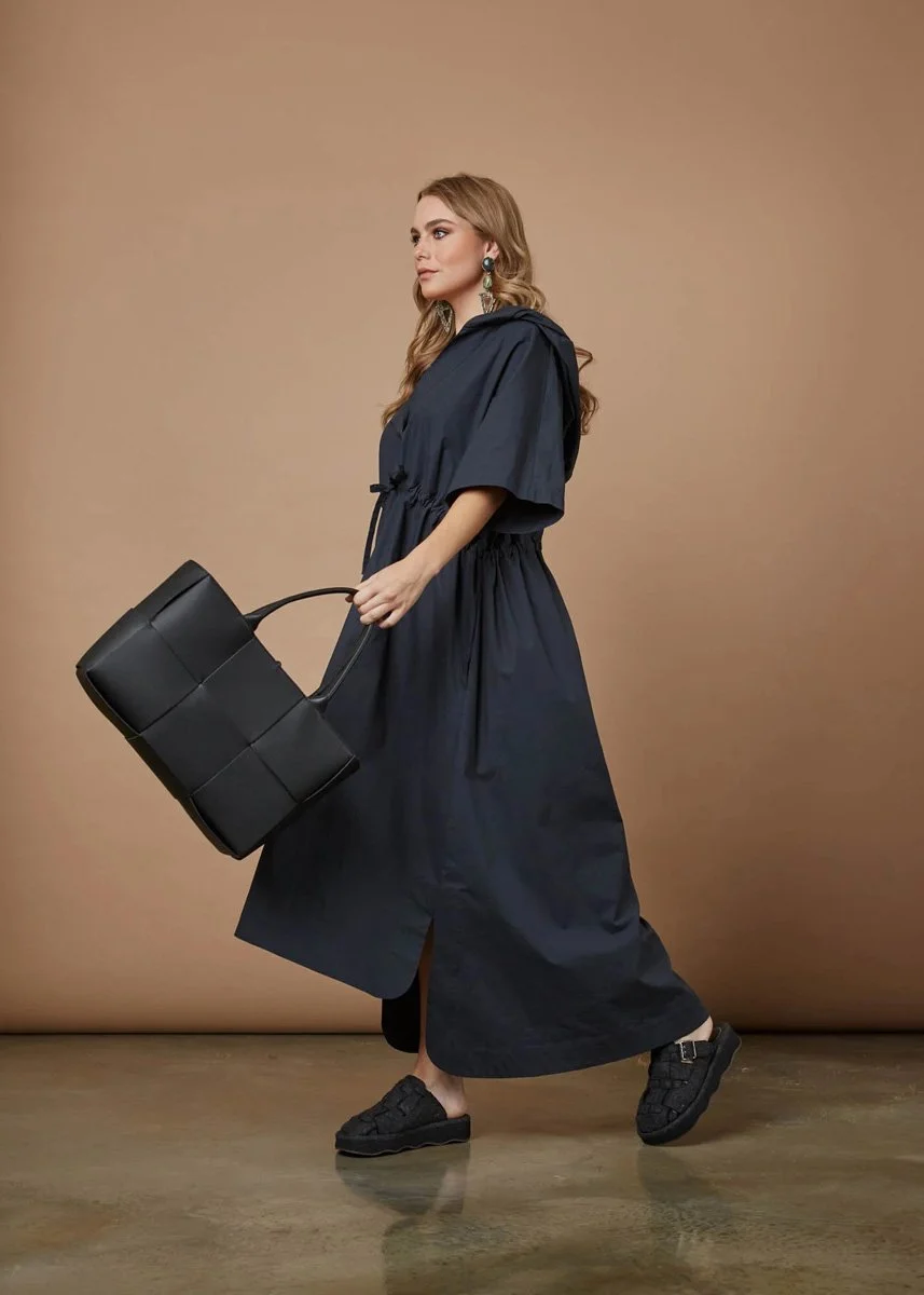 Woman with long blonde hair wearing a navy maxi dress, black sneakers, and large earrings, holding a black handbag, standing against a beige background.