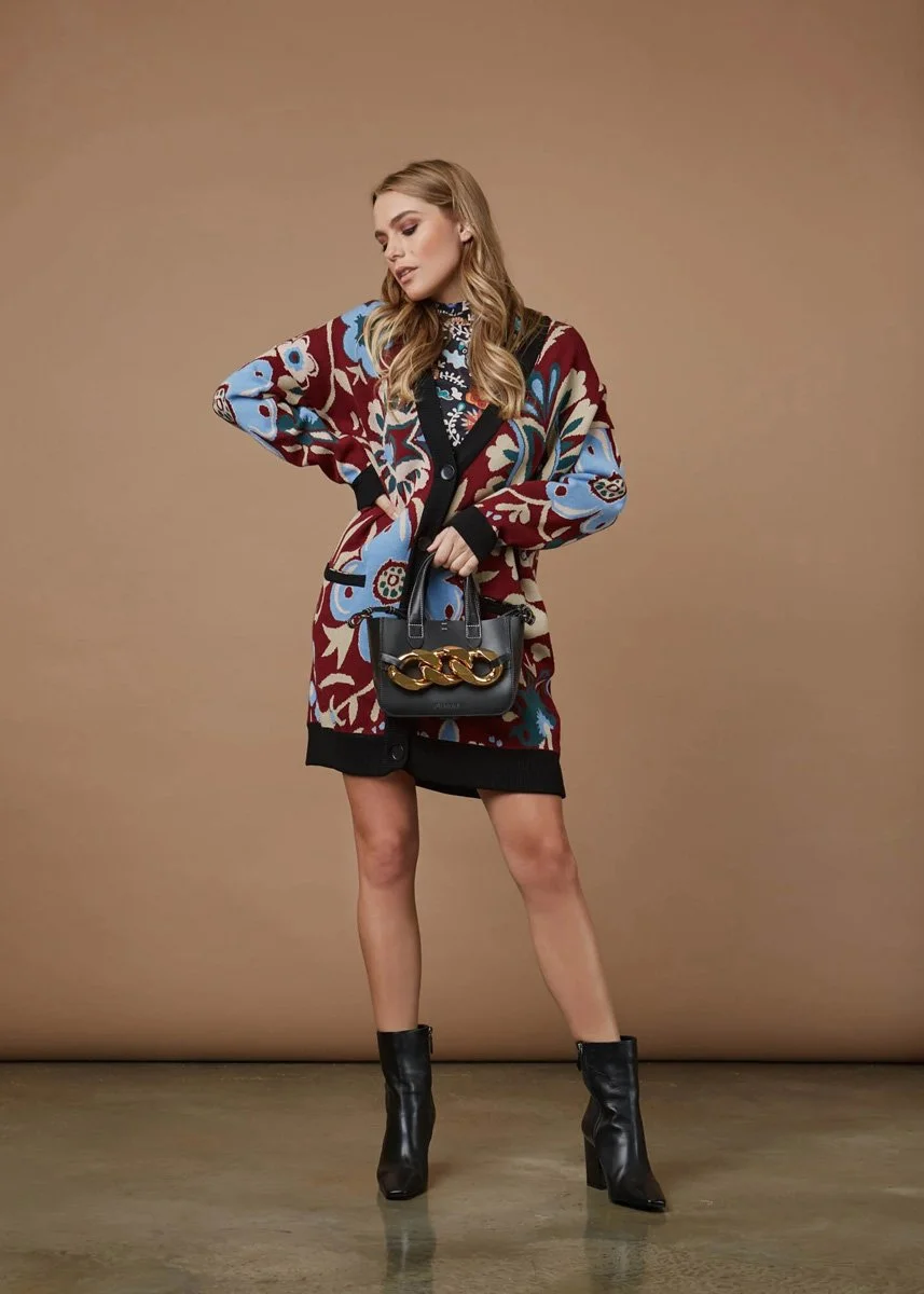 A young woman with long wavy blonde hair, wearing a colorful patterned cardigan and a black dress with a floral design, holding a black handbag with a gold chain, and standing against a plain beige background.