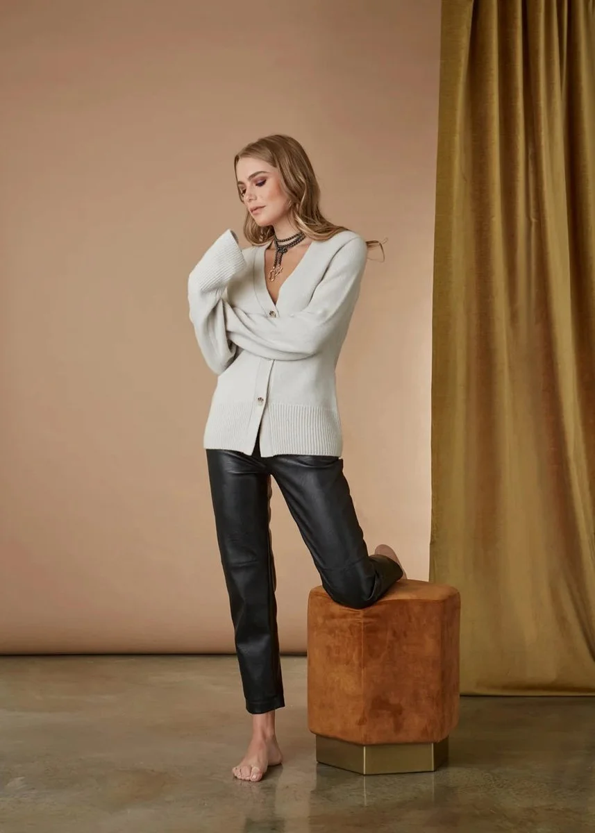A woman with wavy blonde hair wearing a white cardigan, layered necklaces, black leather pants, and no shoes, standing with one knee on a brown velvet stool against a beige background with gold curtains.