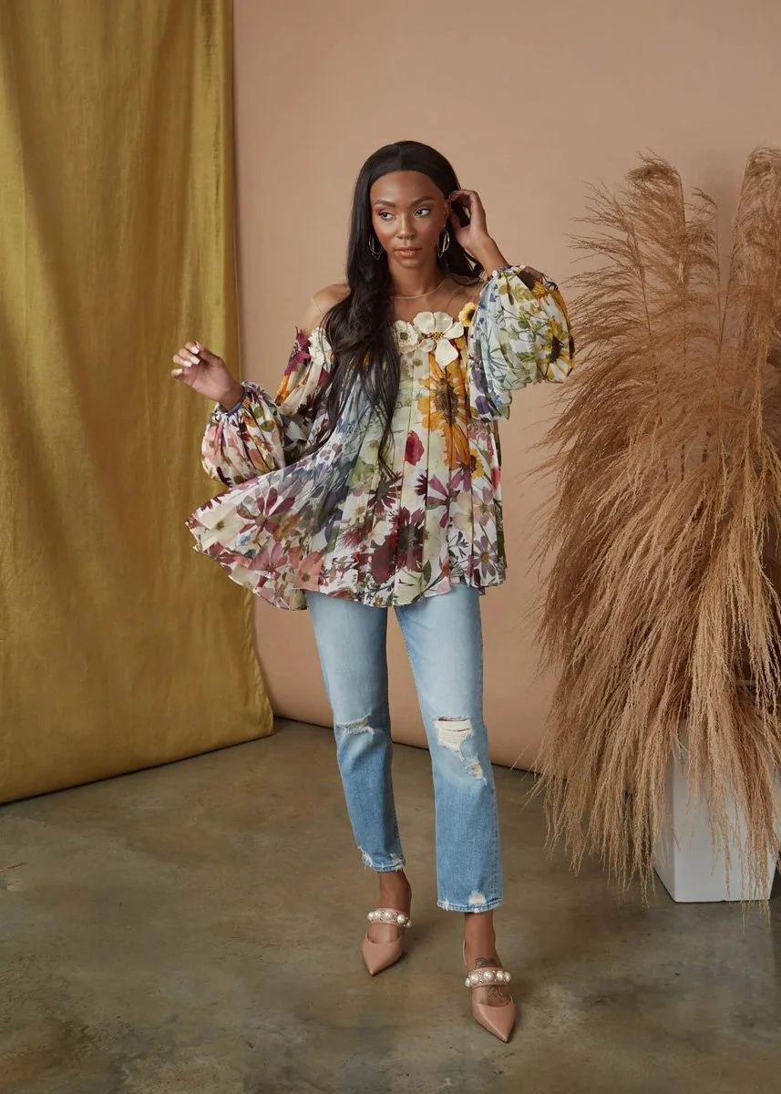 Woman with long black hair wearing a floral off-shoulder blouse with puffy sleeves, ripped jeans, and pink heels standing beside beige curtains and ornamental dried grass in a neutral-colored room.