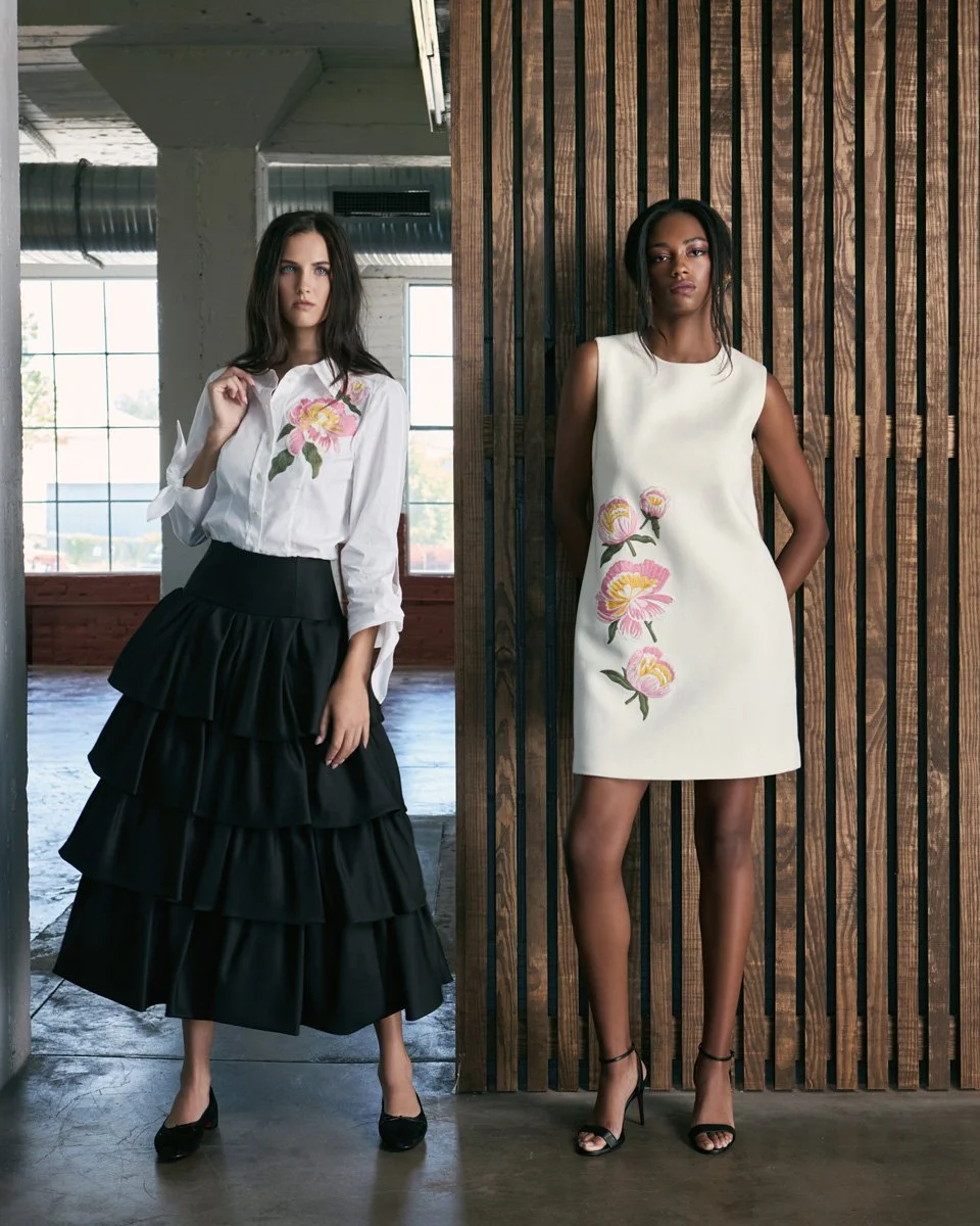 Two women wearing stylish clothing with floral embroidery, standing indoors against a wooden wall and large windows.