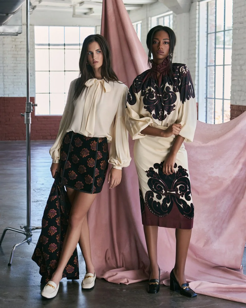 Two women standing in front of a pink fabric backdrop in a spacious, industrial-style room with large windows. They are wearing elegant, patterned dresses with vintage-inspired designs.