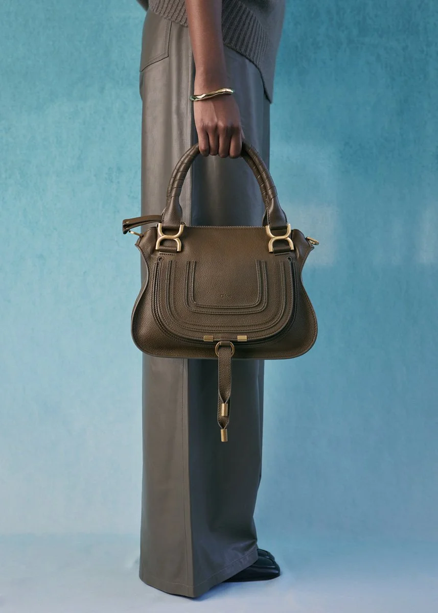 A person holding a brown leather handbag, dressed in gray pants and a dark sweater, against a blue background.