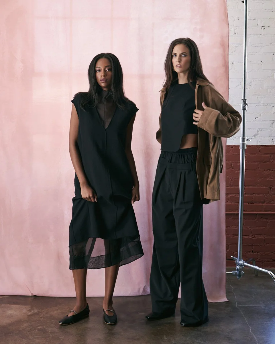 Two women standing in front of a pink backdrop with a brick wall to the right. One woman is wearing a sleeveless black dress with sheer panels, and the other woman is wearing a black top, wide-leg black pants, and a brown jacket, holding the jacket o