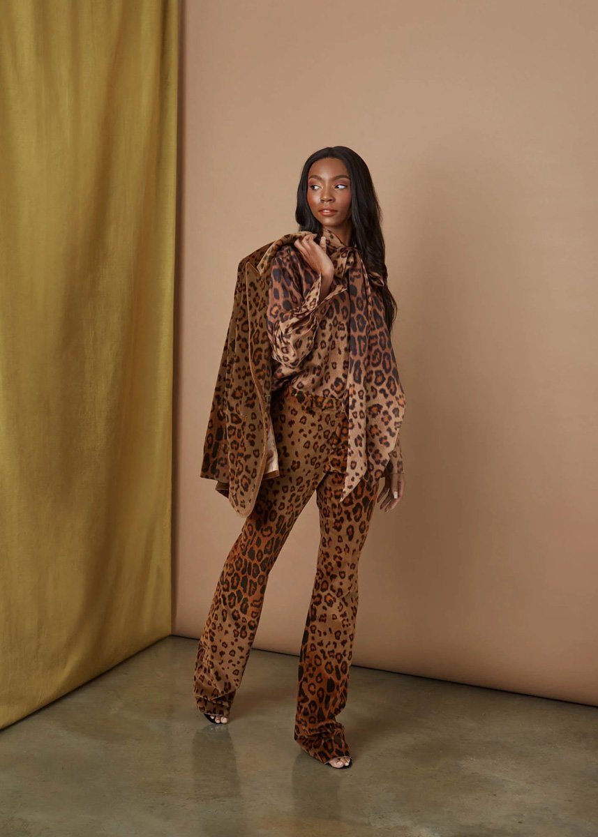 A woman with long dark hair wearing a matching leopard print suit and blazer, standing against a minimal backdrop with beige walls and a golden curtain.