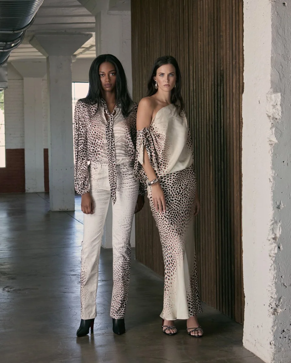 Two women standing side by side in a stylish, modern interior with concrete floors and white brick walls. They are wearing fashionable animal print outfits, one in a cream and black pattern with a blouse and pants, the other in a matching top and pan