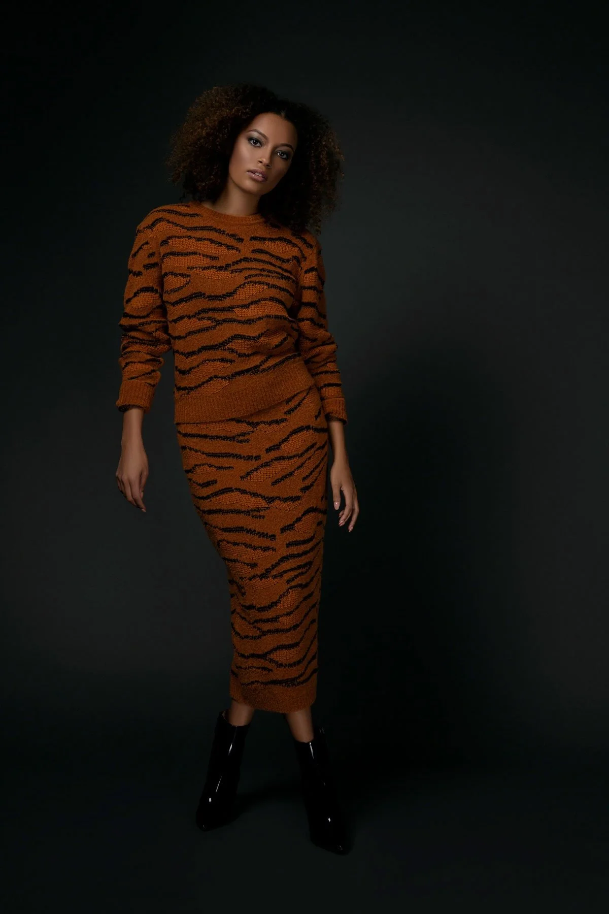 A woman with curly hair wearing a matching orange and black tiger print knit sweater and skirt, paired with black ankle boots, standing against a dark background.