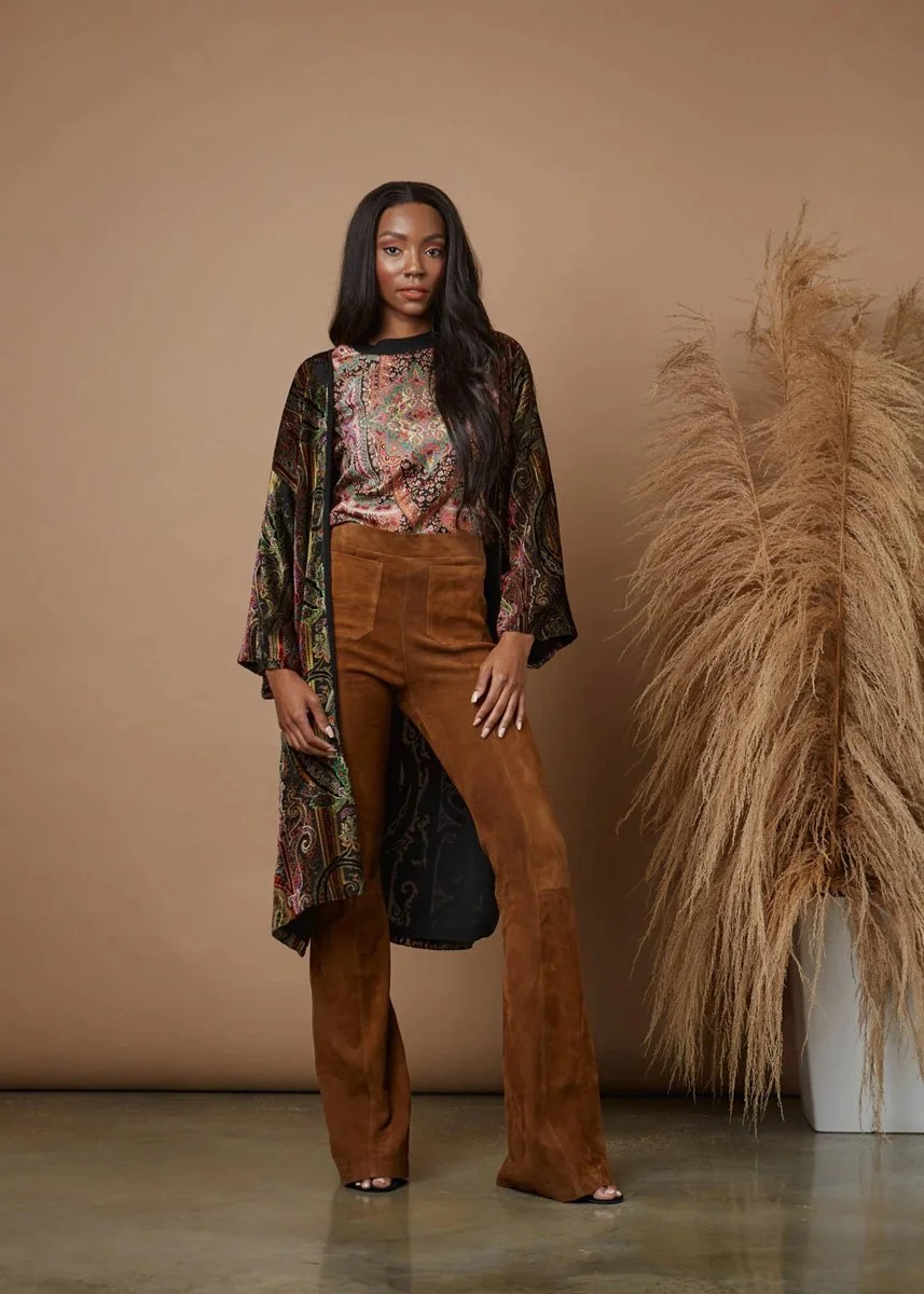 A woman with long black hair wearing high-waisted brown pants, a colorful patterned long-sleeve top, and a flowing patterned outer jacket standing next to a tall vase with large, feathery pampas grass.