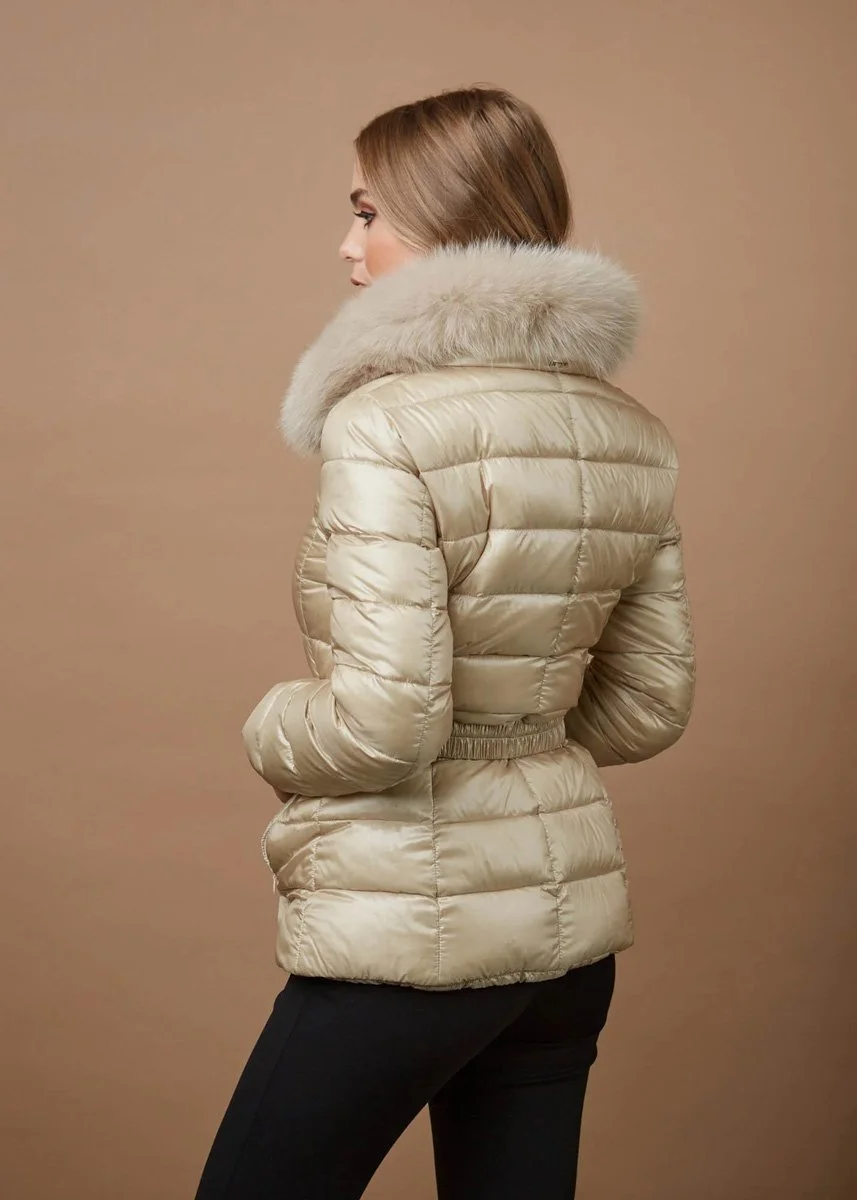 Side view of a woman wearing a beige puffer jacket with a large fur-lined hood, standing against a plain beige background.