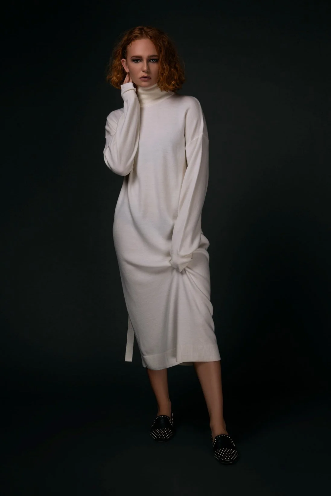 A woman with curly red hair wearing a long, white, turtleneck sweater dress standing against a dark background, with one hand touching her face and the other holding the dress.