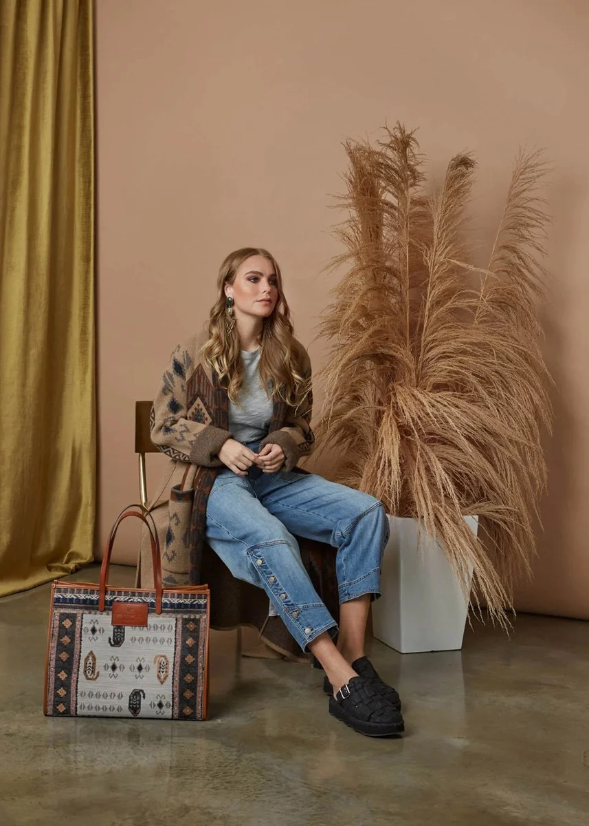 A woman sitting on a wooden chair, wearing a patterned brown coat, a gray shirt, and blue jeans. She has long, wavy blonde hair and is looking to the left. There is a large, decorative tote bag with a southwestern pattern on the floor in front of her