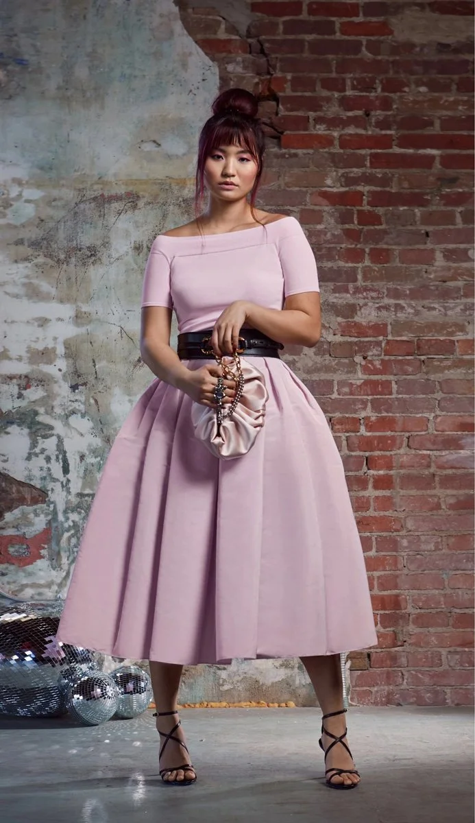 A young woman with a high bun hairstyle standing against a brick and textured wall, wearing a pink dress with a wide black belt, black strappy high heels, and holding a beaded purse in front of her.