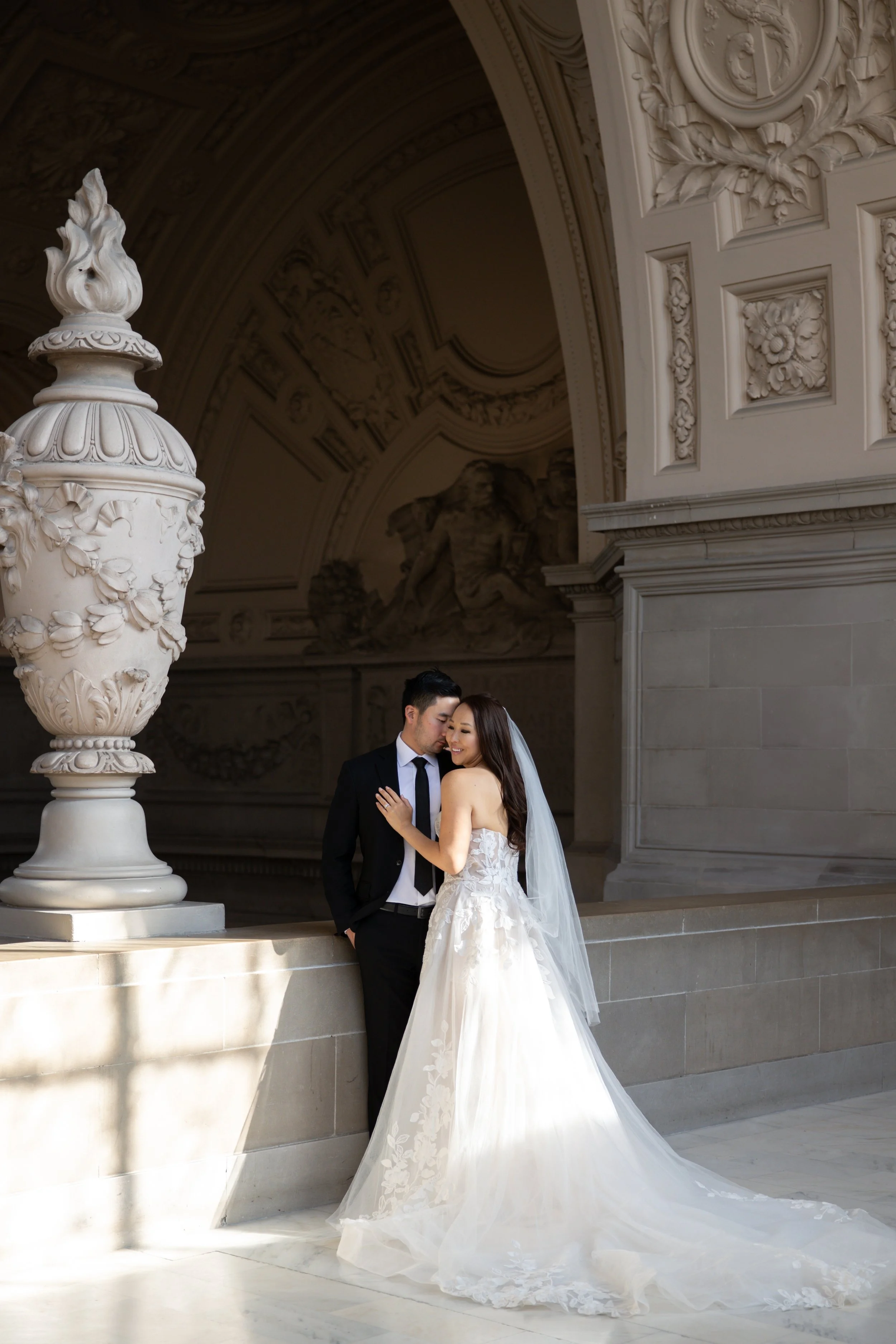 Bride and groom embracing in elegant architectural setting with ornate decor, bride in lace gown and veil, groom in black suit.