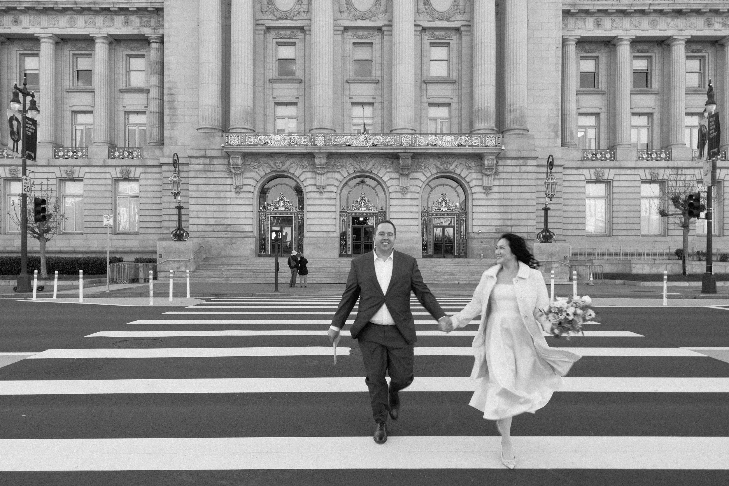 Couple holding hands and crossing the street in front of a historic building, dressed in formal attire, with the woman holding a bouquet.