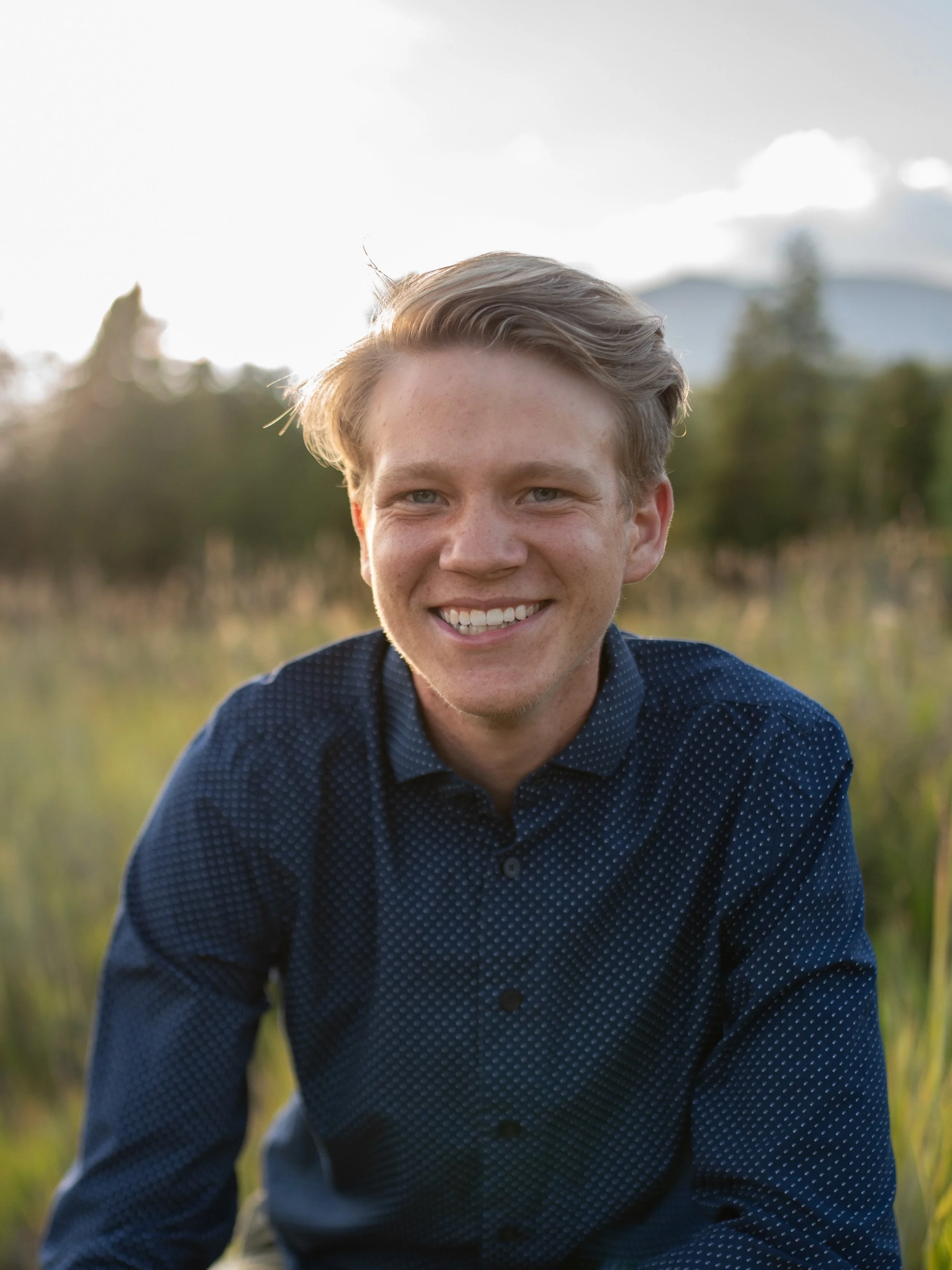High school senior boy smiling in golden hour Colorado mountain meadow with peaks in background — Denver senior portrait photographer Jennie Bennett Photography
