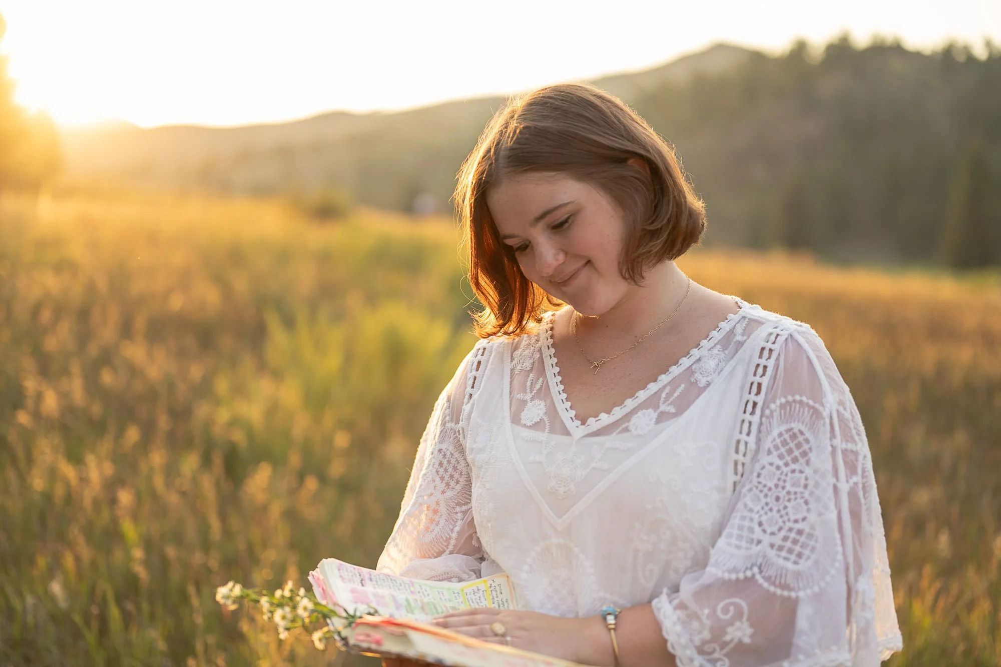 High school senior girl in a white lace top reading in a golden grass meadow at sunset with the Colorado foothills behind her, senior portraits by Jennie Bennett Photography